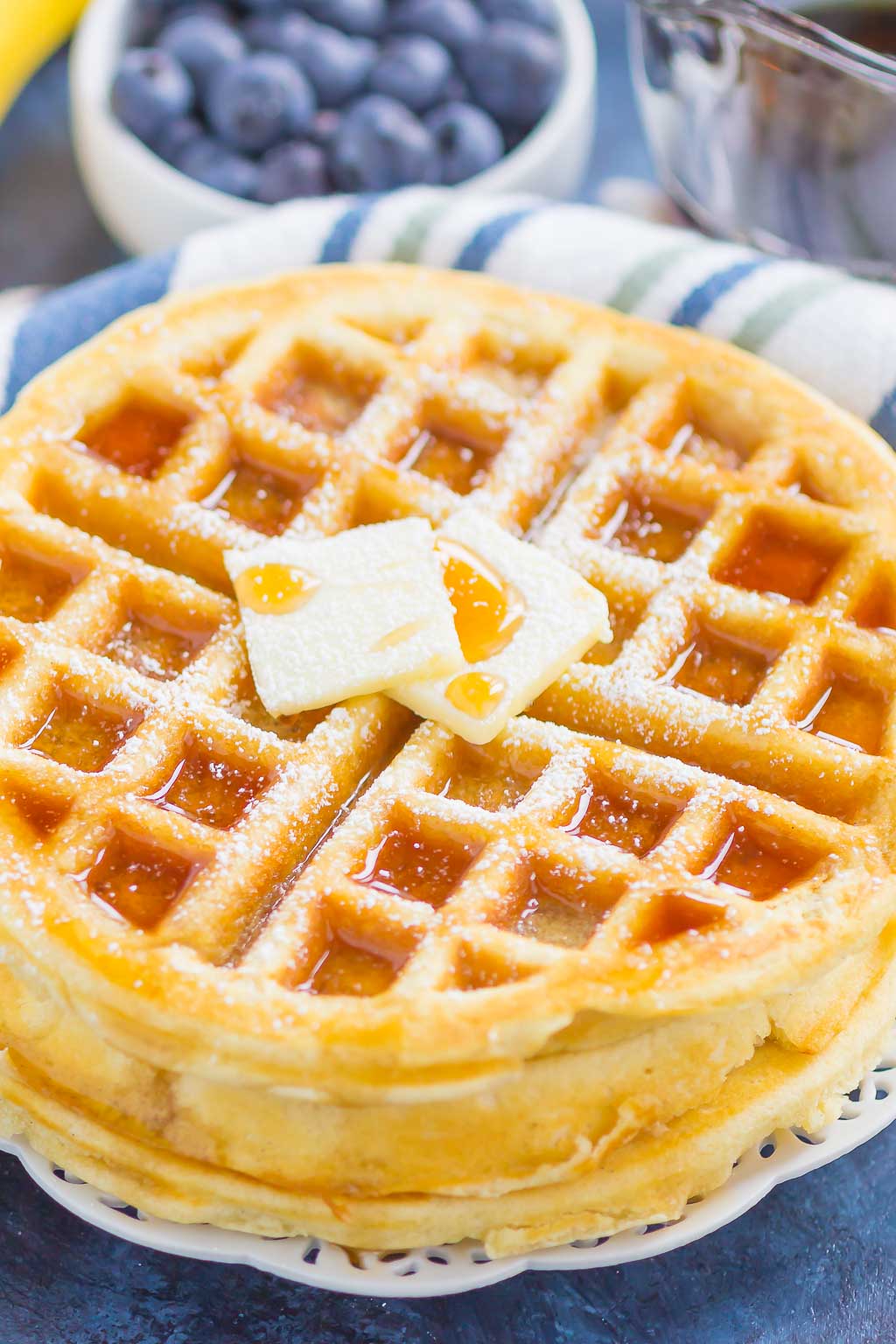 A stack of fluffy waffles topped with butter and maple syrup. A bowl of blueberries rests in the background.